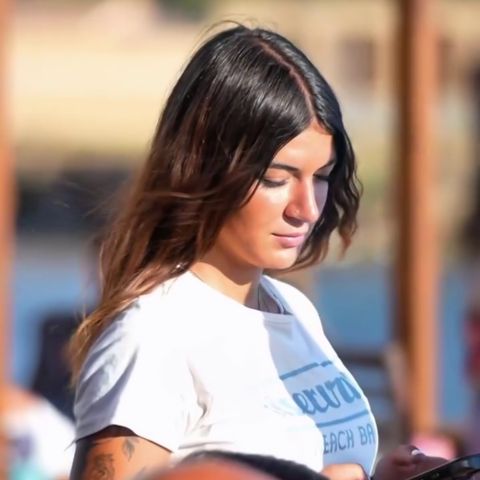 Cameriera tatuata in t-shirt bianca con logo del Riservato Beach bar, guarda il tablet, indossa una divisa personalizzata realizzata da Raimonda Lai.