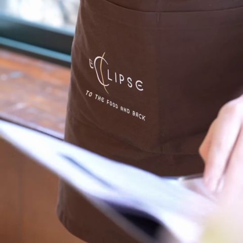 Una persona tiene un menu e indossa un grembiule color cacao con la scritta "Eclipse" su di esso. Il grembiule corto da responsabile di sala.